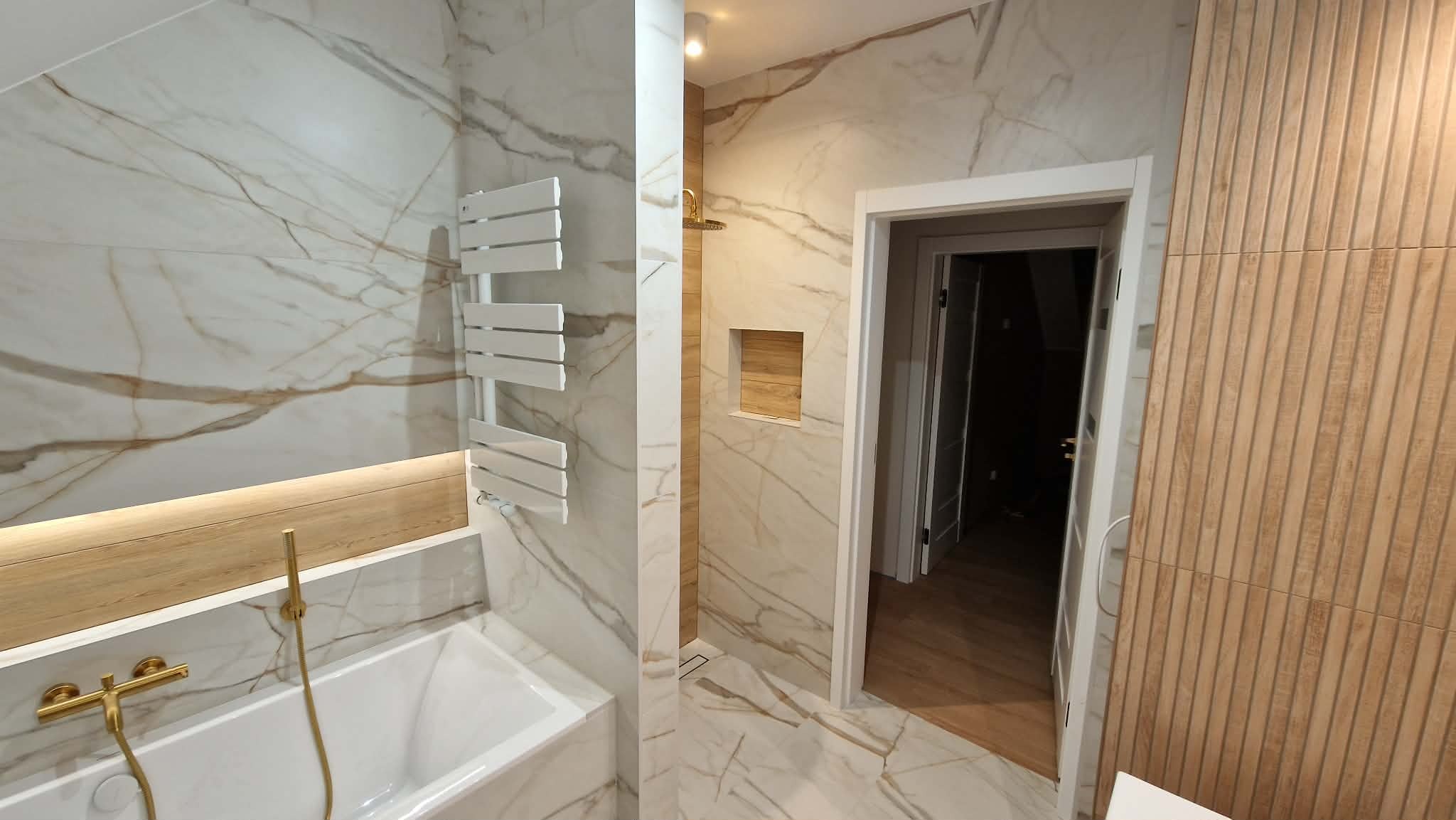 Wykończona łazienka z marmurowymi płytkami, złotą armaturą i grzejnikiem drabinkowym. Widoczne drewniane panele ścienne i oświetlenie LED. Nowoczesny design.