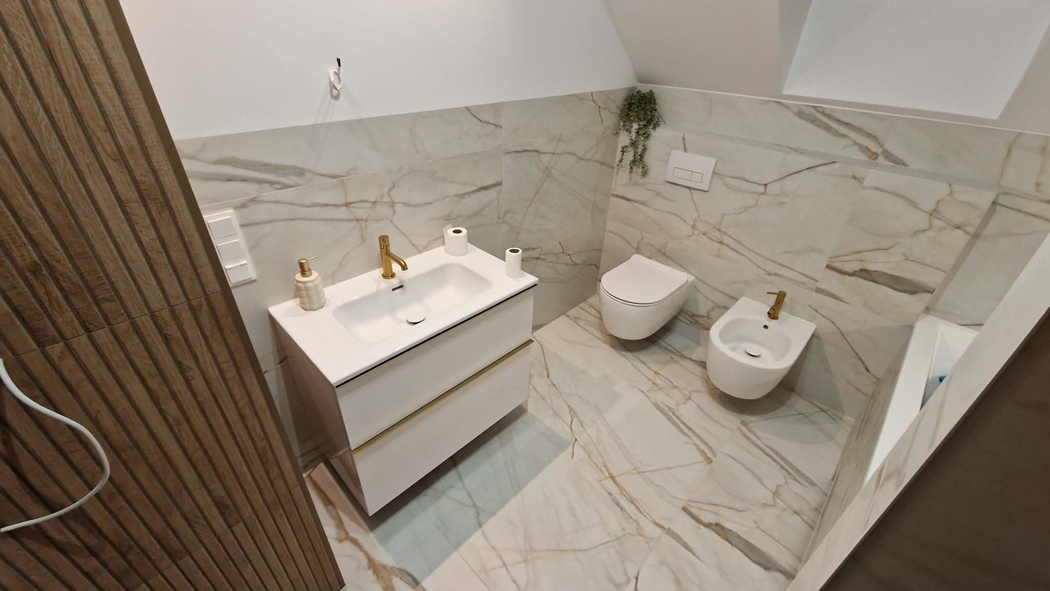 Wykończona łazienka w beżowych płytkach imitujących marmur. Biała szafka z umywalką i złotą armaturą, toaleta i bidet. Dekoracyjne panele ścienne po lewej.