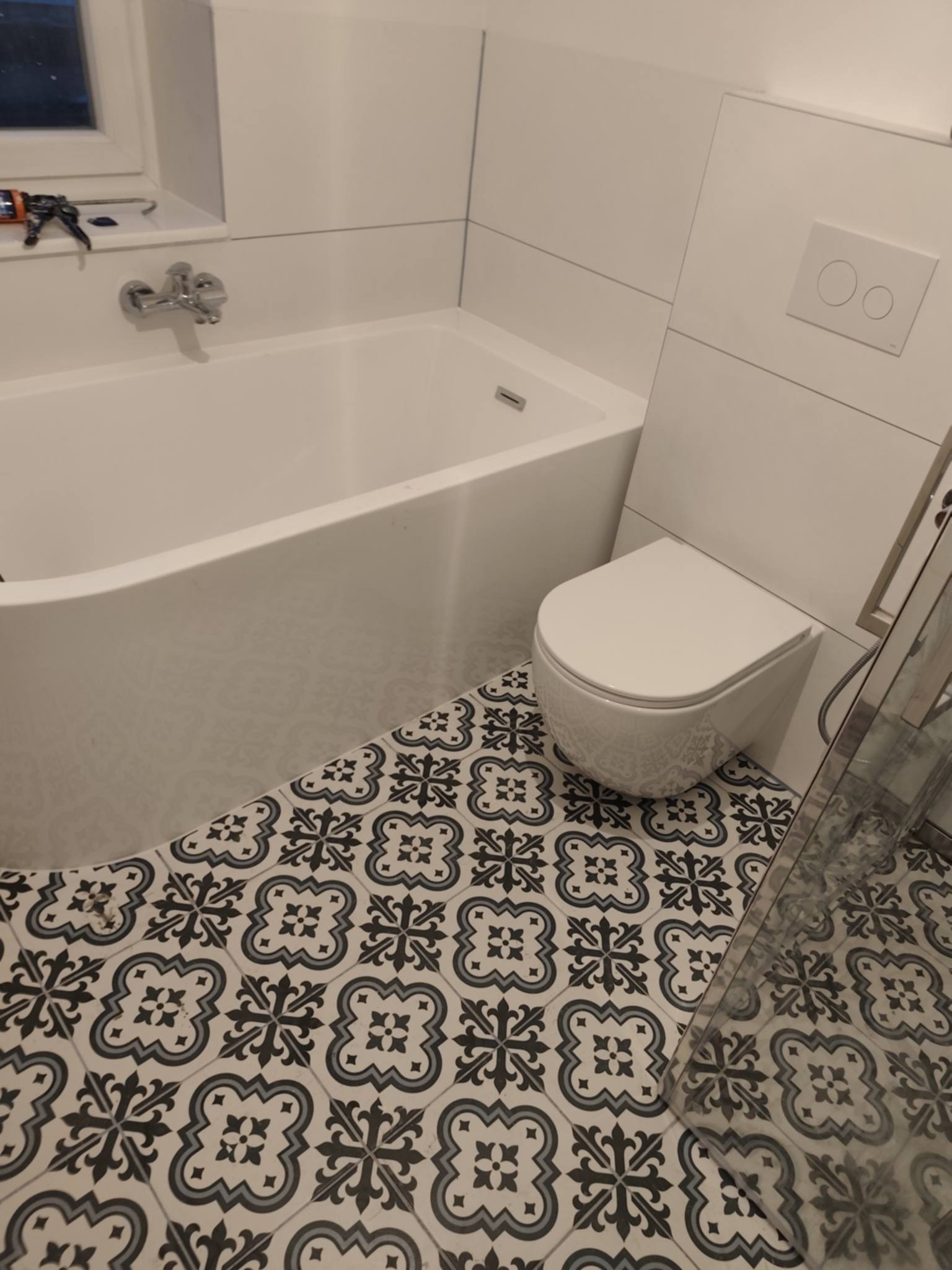 Biała wanna i toaleta na podłodze z wzorzystych, czarno-białych płytek w wykończonej łazience. Widoczny fragment kabiny prysznicowej i narzędzia na parapecie.