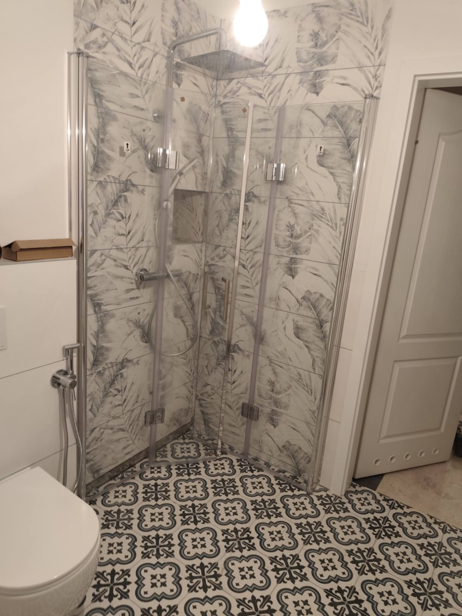 Wykończona kabina prysznicowa z wzorzystymi płytkami na ścianach i podłodze. Widoczna toaleta i fragment drzwi. Dekoracyjne płytki z motywem roślinnym i geometrycznym.
