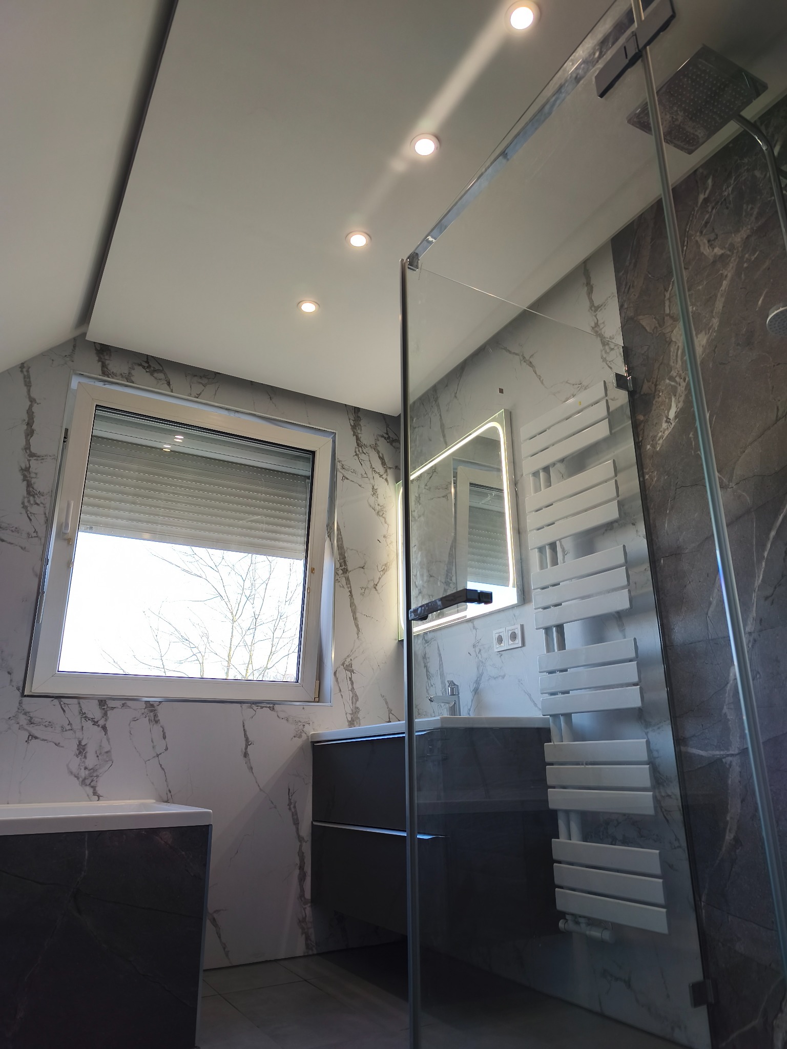 Nowoczesna łazienka na poddaszu z marmurowymi płytkami, kabiną prysznicową typu walk-in, grzejnikiem drabinkowym i oknem z roletami zewnętrznymi.