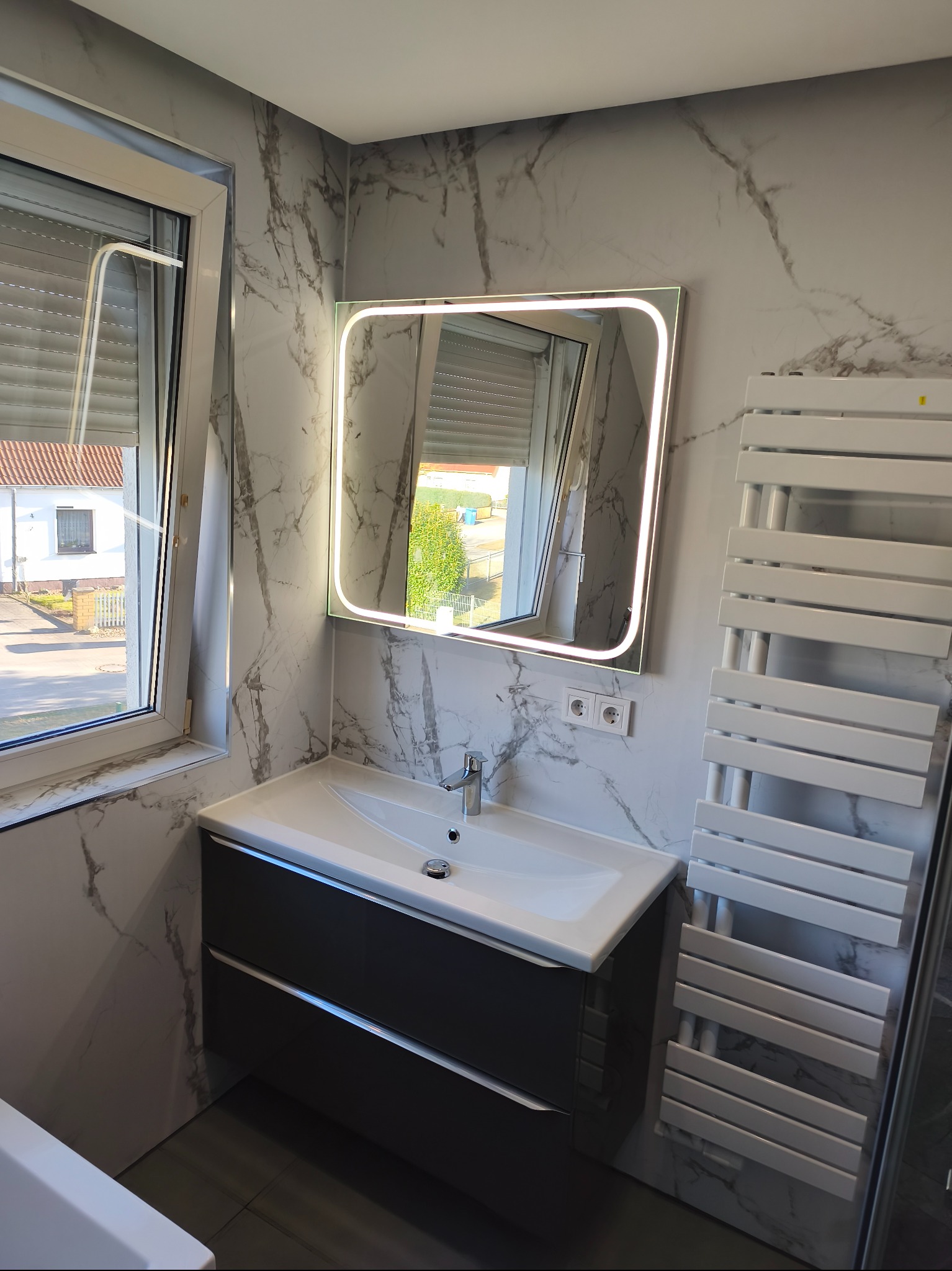 Nowoczesna łazienka z umywalką w szafce, podświetlanym lustrem i grzejnikiem drabinkowym na tle marmurowych płytek. Widok z okna na dom.