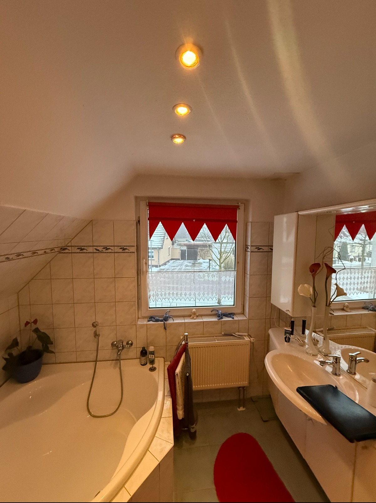 Jasna łazienka z oknem ozdobionym czerwoną roletą, białą wanną i umywalką. Na ścianach beżowe płytki. Widoczne grzejnik i dekoracje okienne.