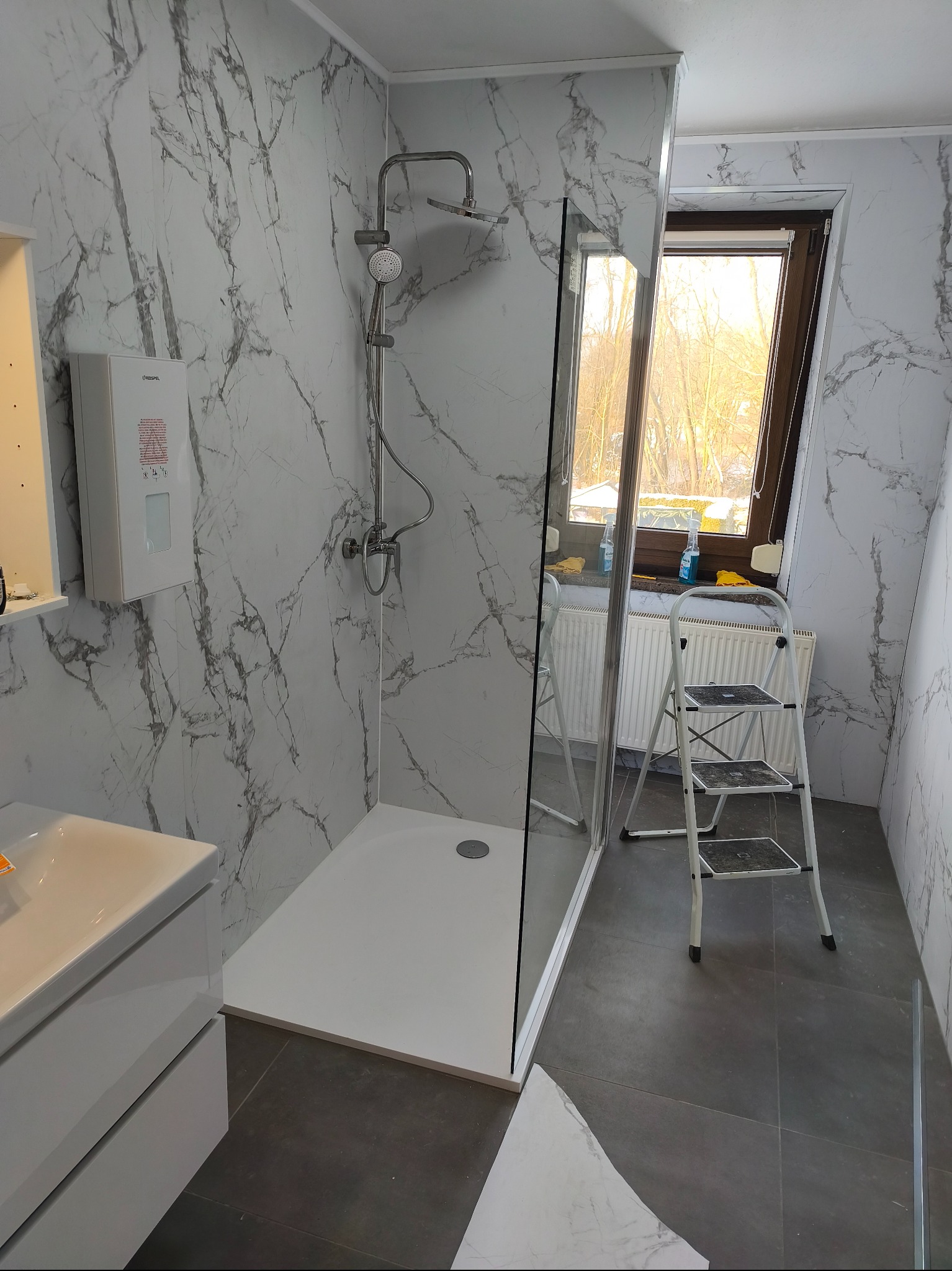 Wykończona łazienka z kabiną prysznicową typu walk-in, białą armaturą i panelami ściennymi imitującymi marmur. Widoczna drabina malarska i okno dachowe.