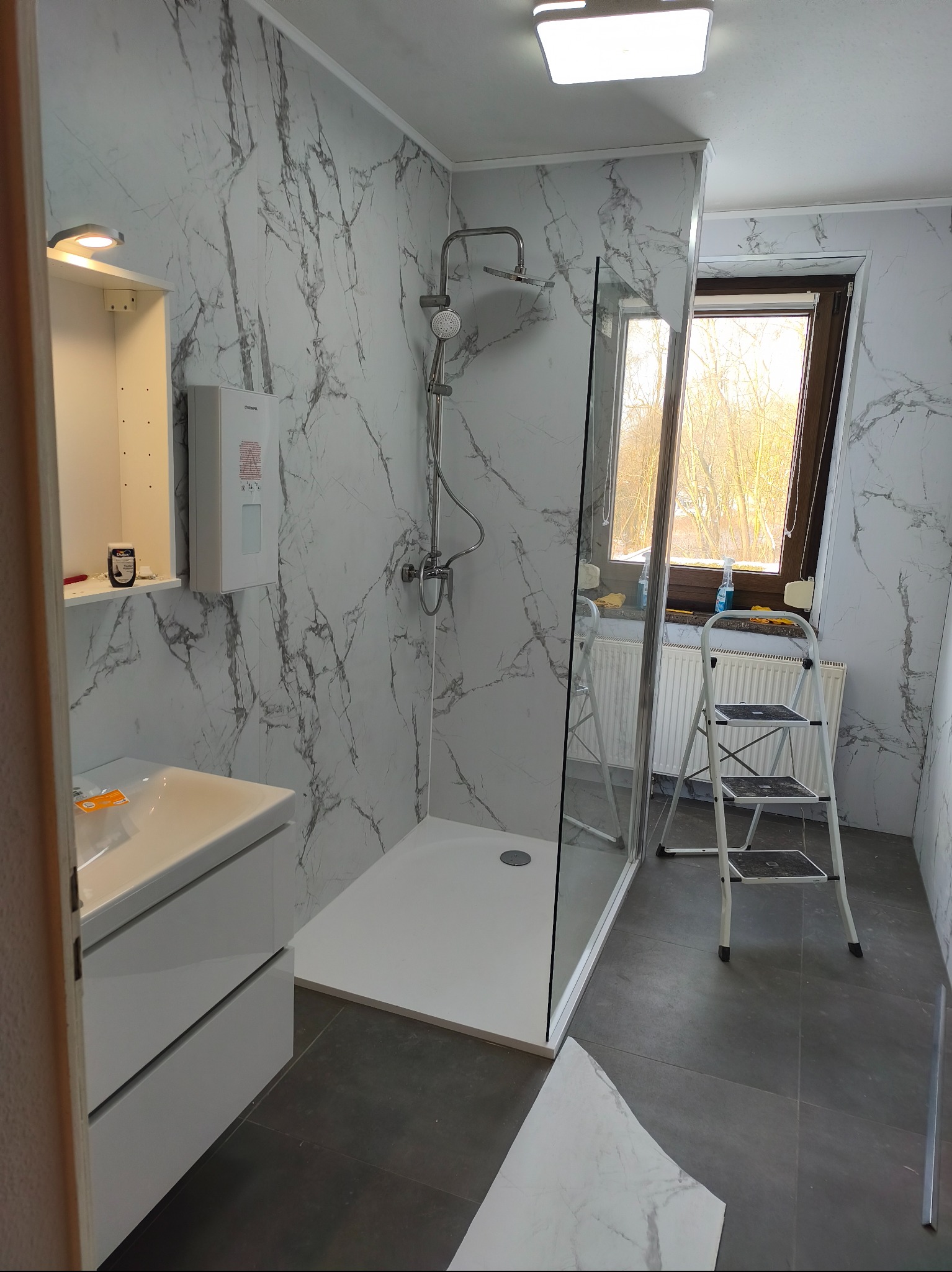 Wykończona łazienka z kabiną prysznicową, umywalką z szafką i grzejnikiem pod oknem. Ściany wyłożone płytami imitującymi marmur. Na podłodze szare płytki.