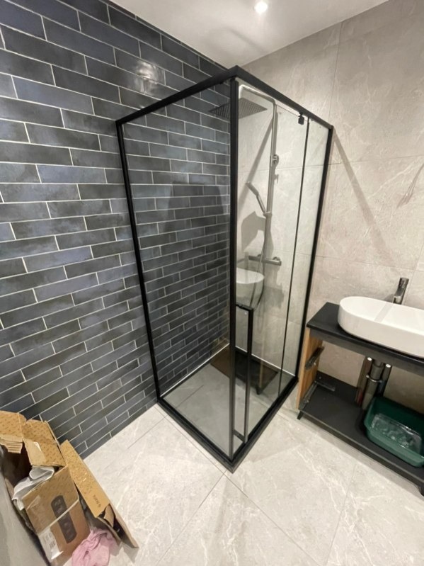 Nowoczesna kabina prysznicowa z czarną ramą i deszczownicą, obok umywalka na szafce. Ściany wyłożone szarą cegłą i płytkami imitującymi marmur. Na podłodze jasne płytki.