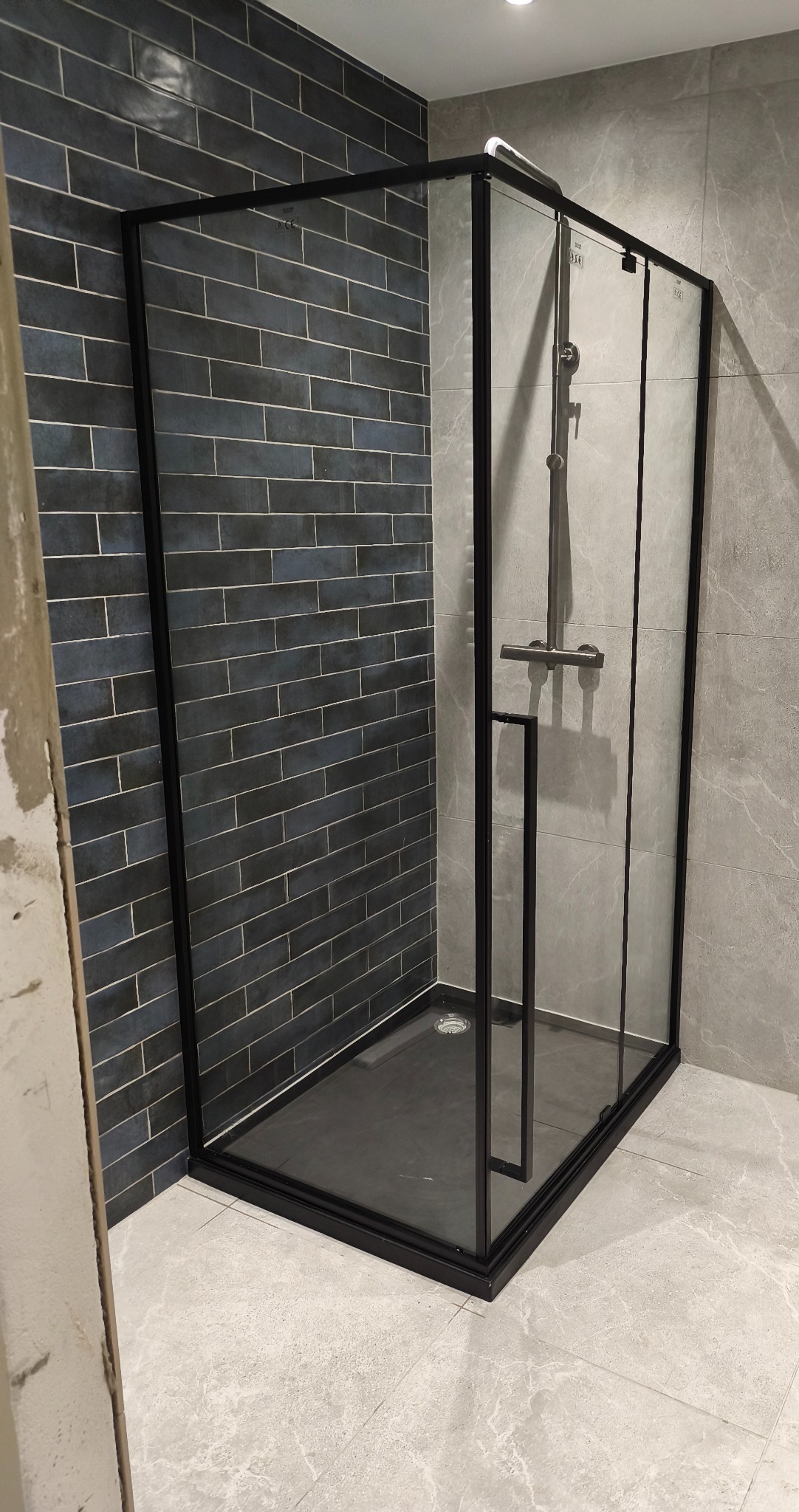 Nowoczesna kabina prysznicowa z czarnymi profilami i ciemnymi płytkami imitującymi cegłę, uzupełniona szarymi płytkami na ścianie i podłodze.
