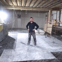 Atamanchuk Denys - Remont wnętrza: glazurnik stoi na ułożonej, czarno-szarej podłodze z płytek. Widoczne profile sufitowe, ściany przygotowane do gładzi, rusztowanie i płyty. W tle drzwi.