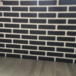 Atamanchuk Denys - Ściana z czarnych, matowych cegiełek na białej fudze, ułożonych rzędami. Na dole widoczna podłoga z paneli imitujących drewno. Minimalistyczny design.
