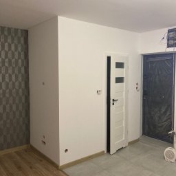 Atamanchuk Denys - Wnętrze po remoncie: białe ściany, wzorzysta tapeta, drewniana podłoga przechodząca w szare płytki, białe drzwi z przeszkleniami i zabezpieczone folią drzwi wejściowe.