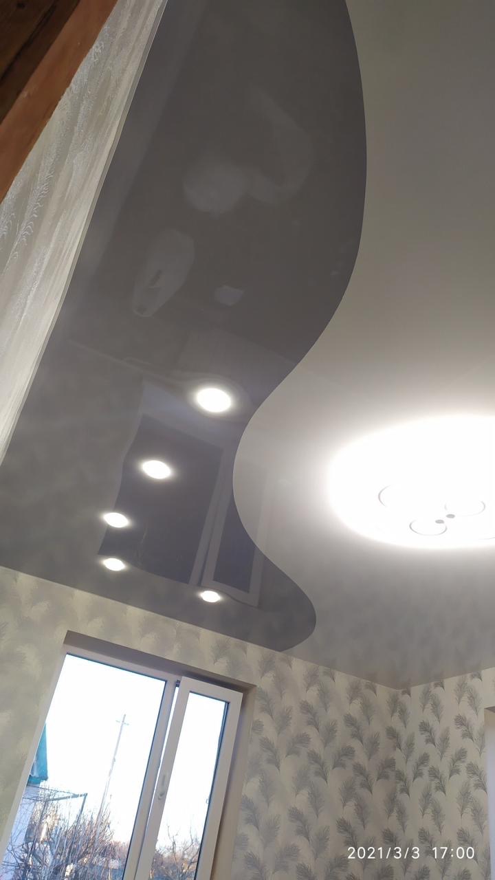 Dwupoziomowy sufit podwieszany z połyskiem, oświetlony punktowo i centralnie, w pokoju z wzorzystą tapetą i oknem. Widok z dołu, ukośny kadr.