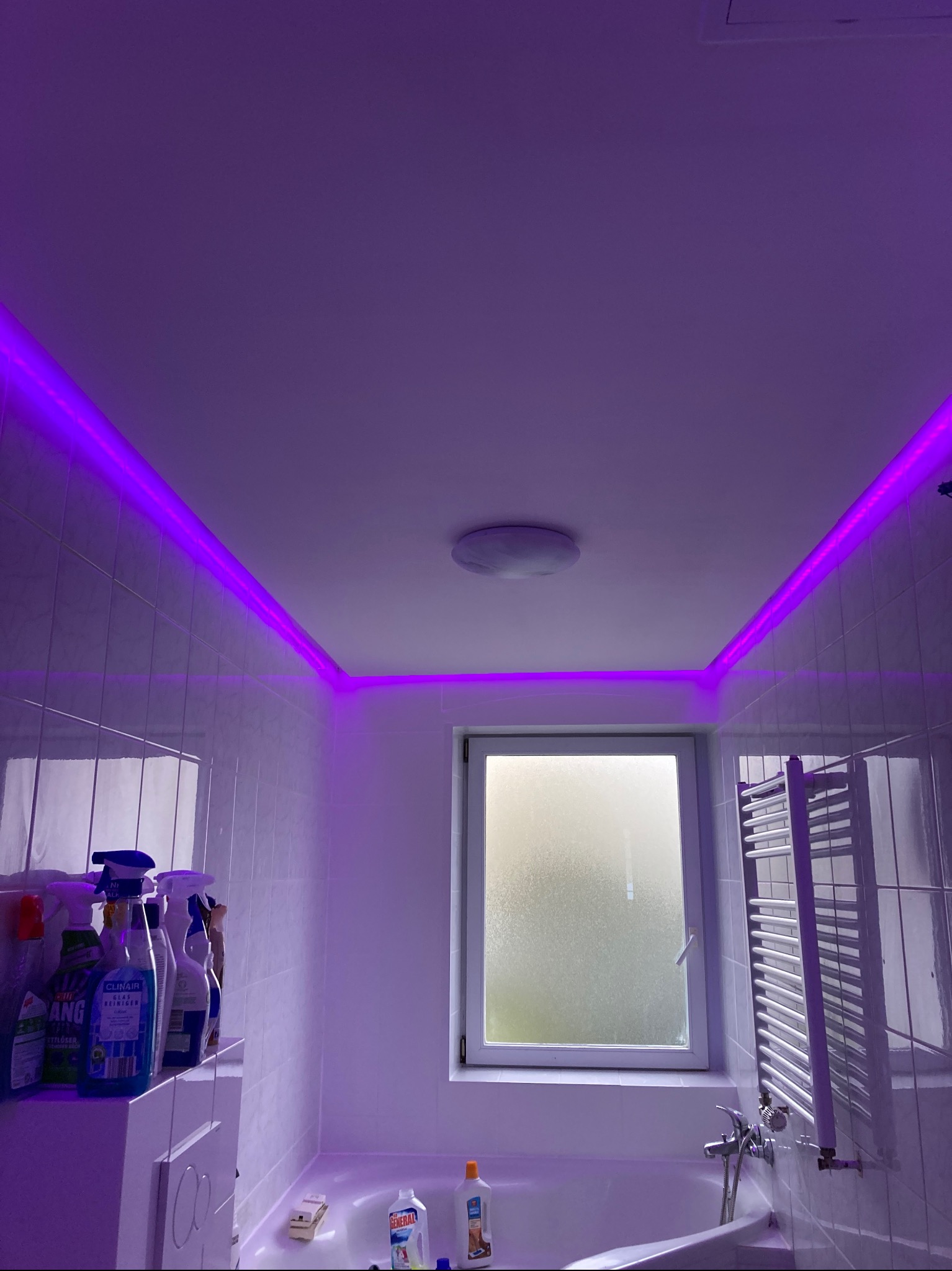 Nowoczesna łazienka z białymi płytkami, oświetlona fioletowymi listwami LED pod sufitem. Widoczne okno, grzejnik drabinkowy i środki czystości.