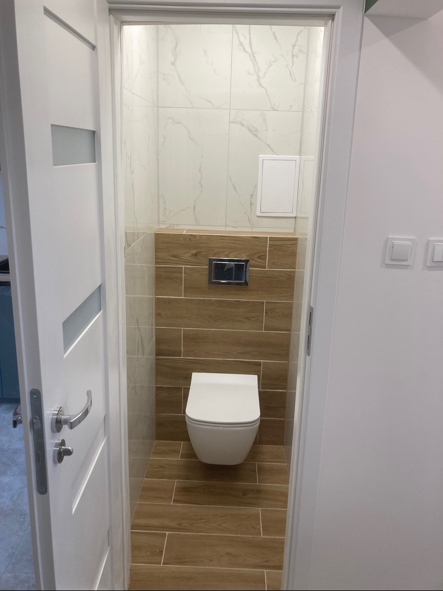 Nowoczesna toaleta w odcieniach bieli i brązu, z podwieszaną miską WC i płytkami imitującymi drewno, widoczna zza uchylonych białych drzwi z przeszkleniem.