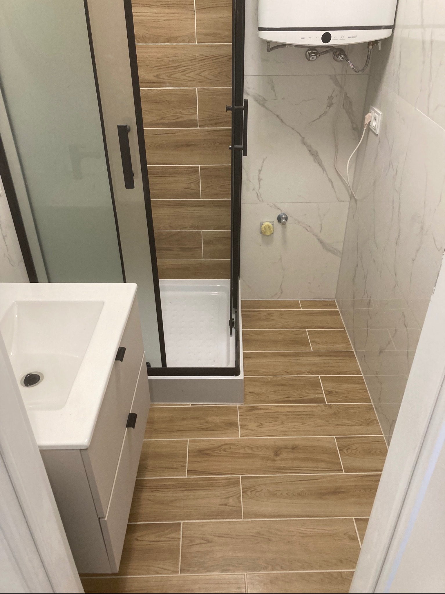 Nowoczesna łazienka po remoncie: kabina prysznicowa z czarnymi ramami, umywalka z szafką, płytki imitujące drewno na podłodze i ścianie, marmurowe płytki na ścianie.