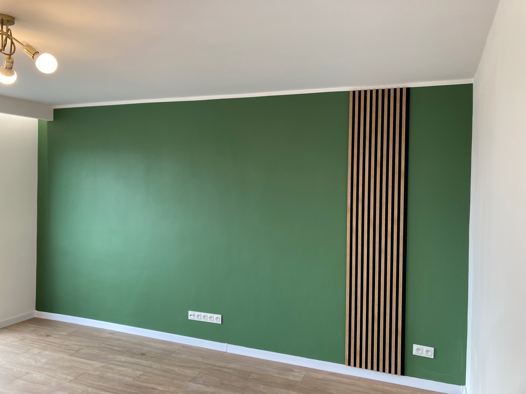 Zielona ściana z drewnianymi lamelami w pionowym układzie. Widoczne gniazdka elektryczne i fragment podłogi z paneli. Oświetlenie sufitowe.