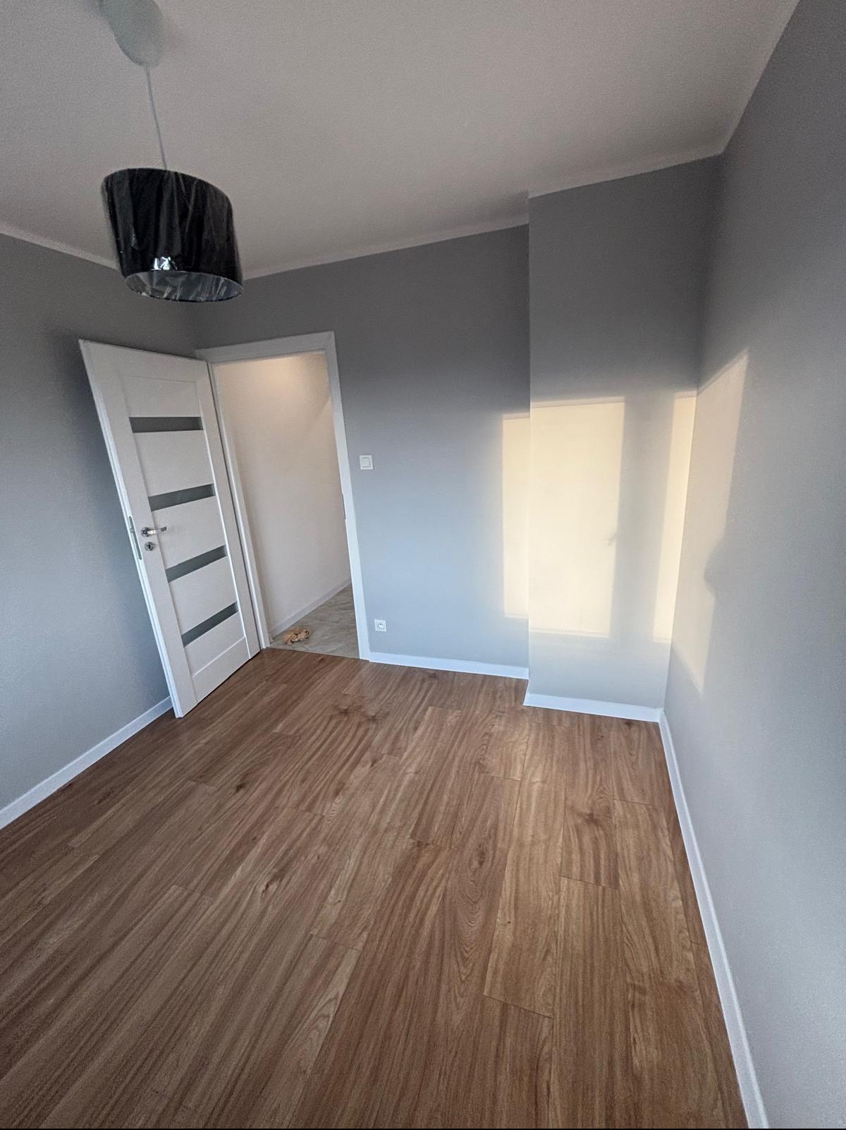 Pomalowane na szaro wnętrze pokoju z drewnianą podłogą, białe listwy przypodłogowe i drzwi z przeszkleniami. Widoczna lampa z czarnym abażurem.