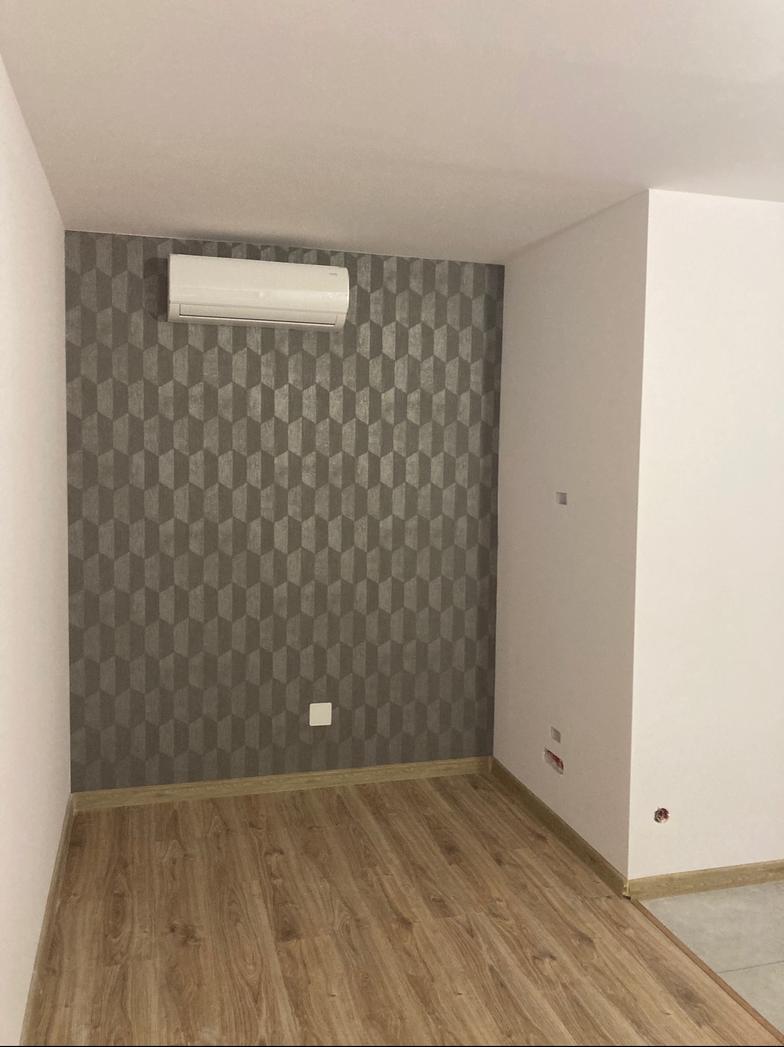 Pokój po remoncie: drewniana podłoga, ściana z wzorzystą tapetą i klimatyzator. Widoczne gniazdka elektryczne. Minimalistyczny design wnętrza.
