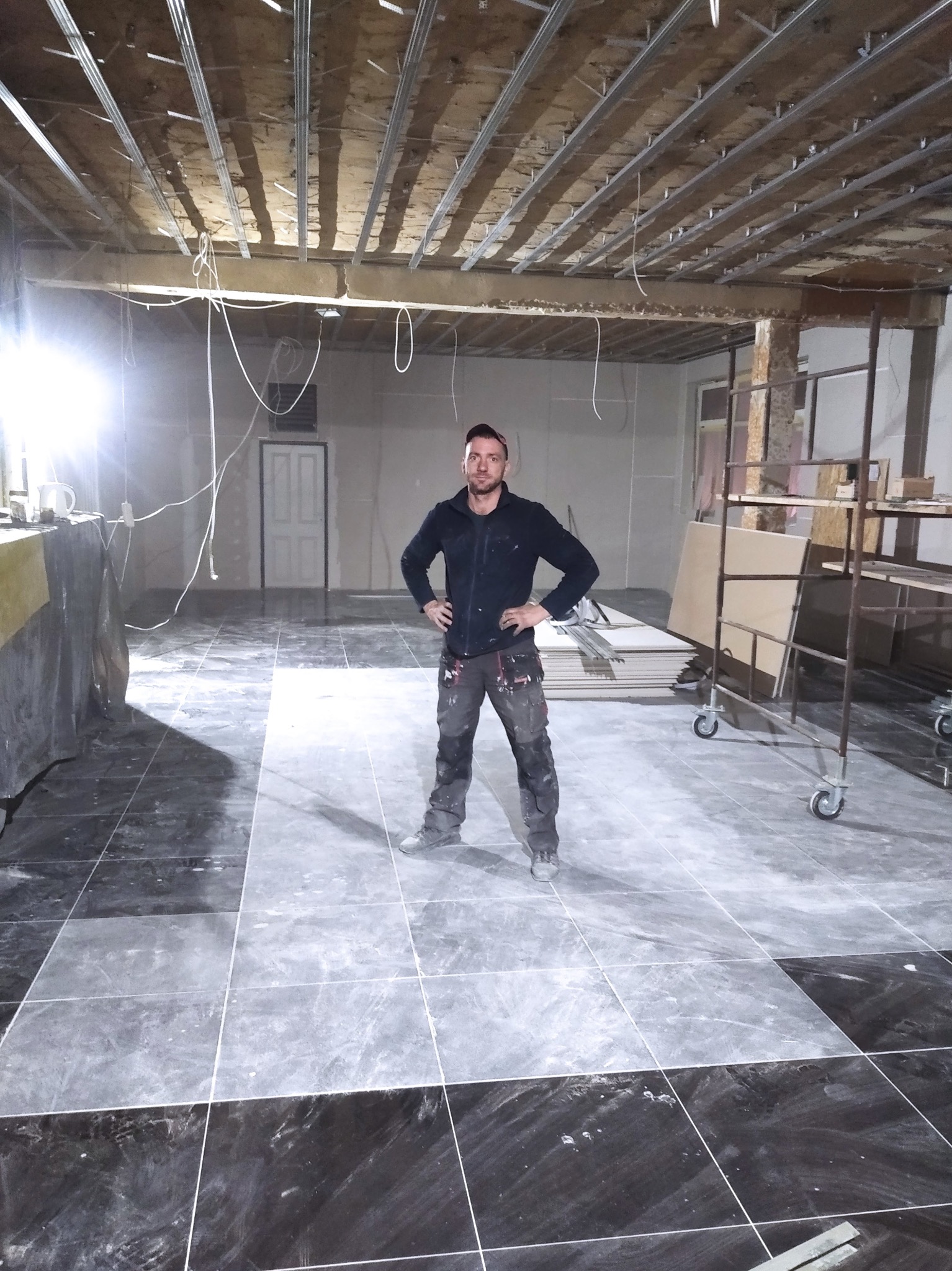 Remont wnętrza: glazurnik stoi na ułożonej, czarno-szarej podłodze z płytek. Widoczne profile sufitowe, ściany przygotowane do gładzi, rusztowanie i płyty. W tle drzwi.