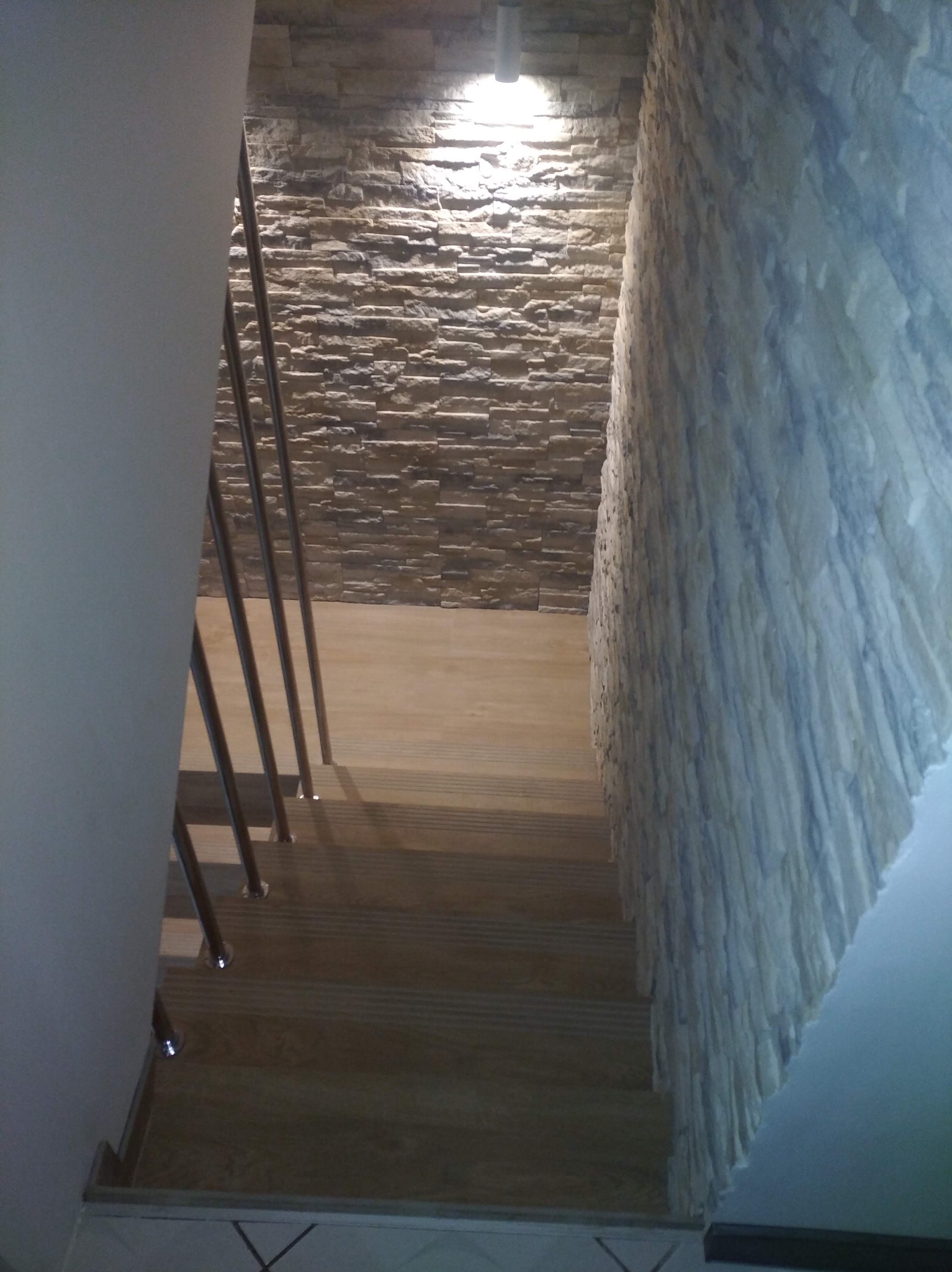 Wykończenie schodów z kamienną okładziną ściany i drewnianymi stopniami. Metalowa barierka. Oświetlenie punktowe. Widok z góry.