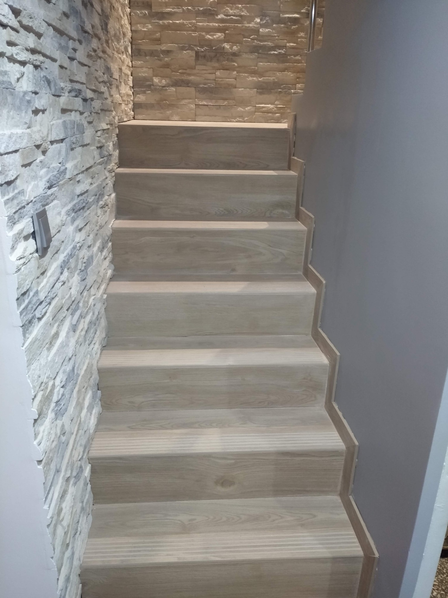 Schody wyłożone panelami imitującymi drewno, z dekoracyjną ścianą z kamienia i gładką, szarą ścianą. Ujęcie z dołu, podkreślające wykończenie.