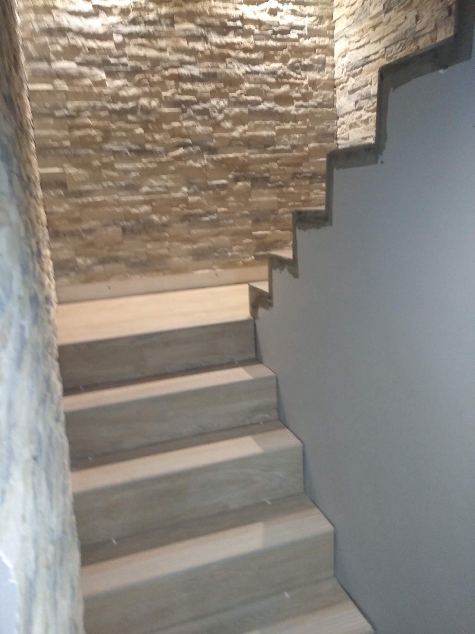 Schody wykończone płytkami imitującymi drewno, ściana z dekoracyjnej cegły w odcieniach beżu i szarości, gładka szara ściana. Nowoczesne wykończenie.