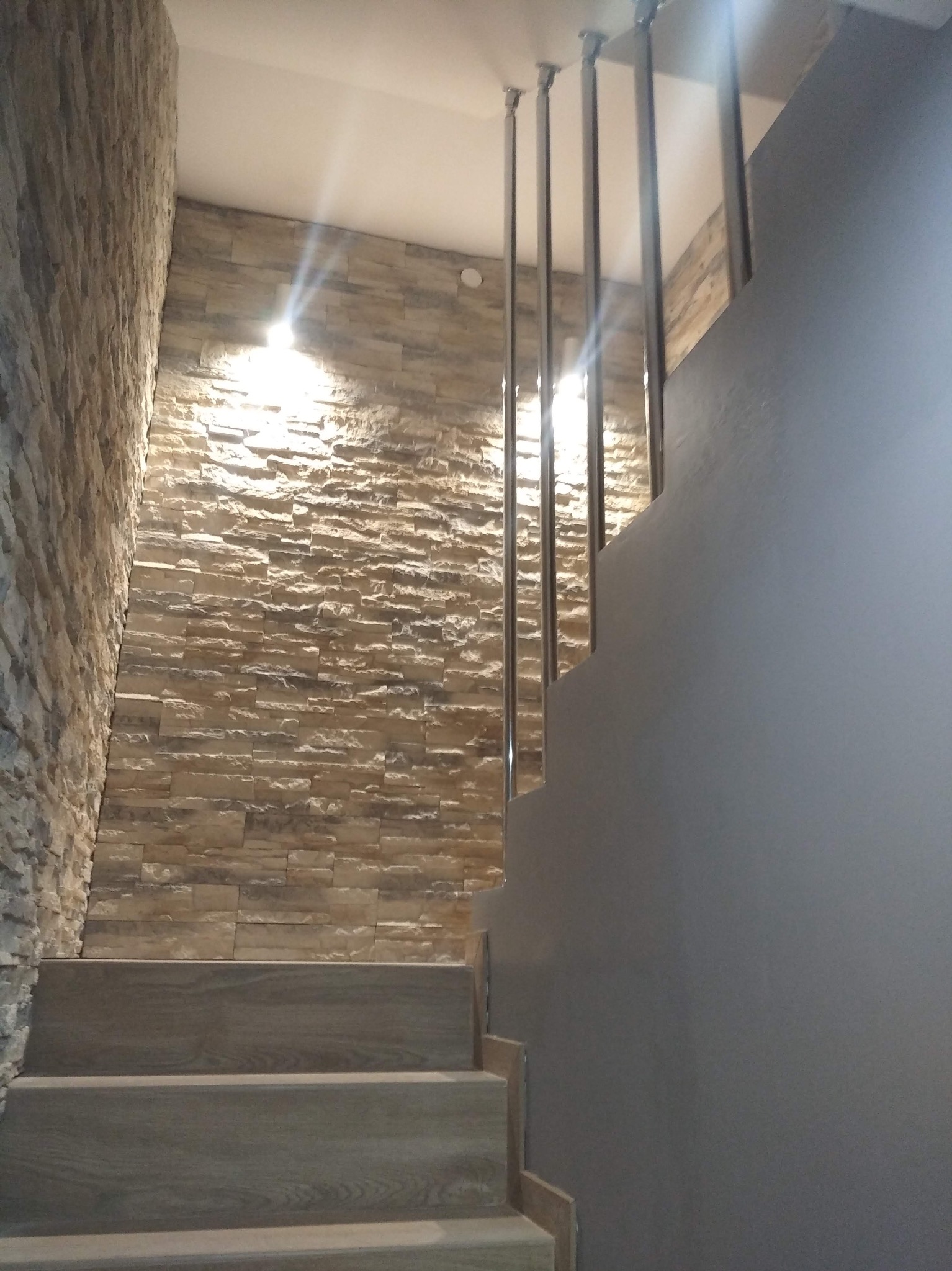 Schody z okładziną kamienną na ścianie i metalową balustradą. Szara ściana po prawej stronie. Oświetlenie punktowe podkreśla fakturę kamienia.
