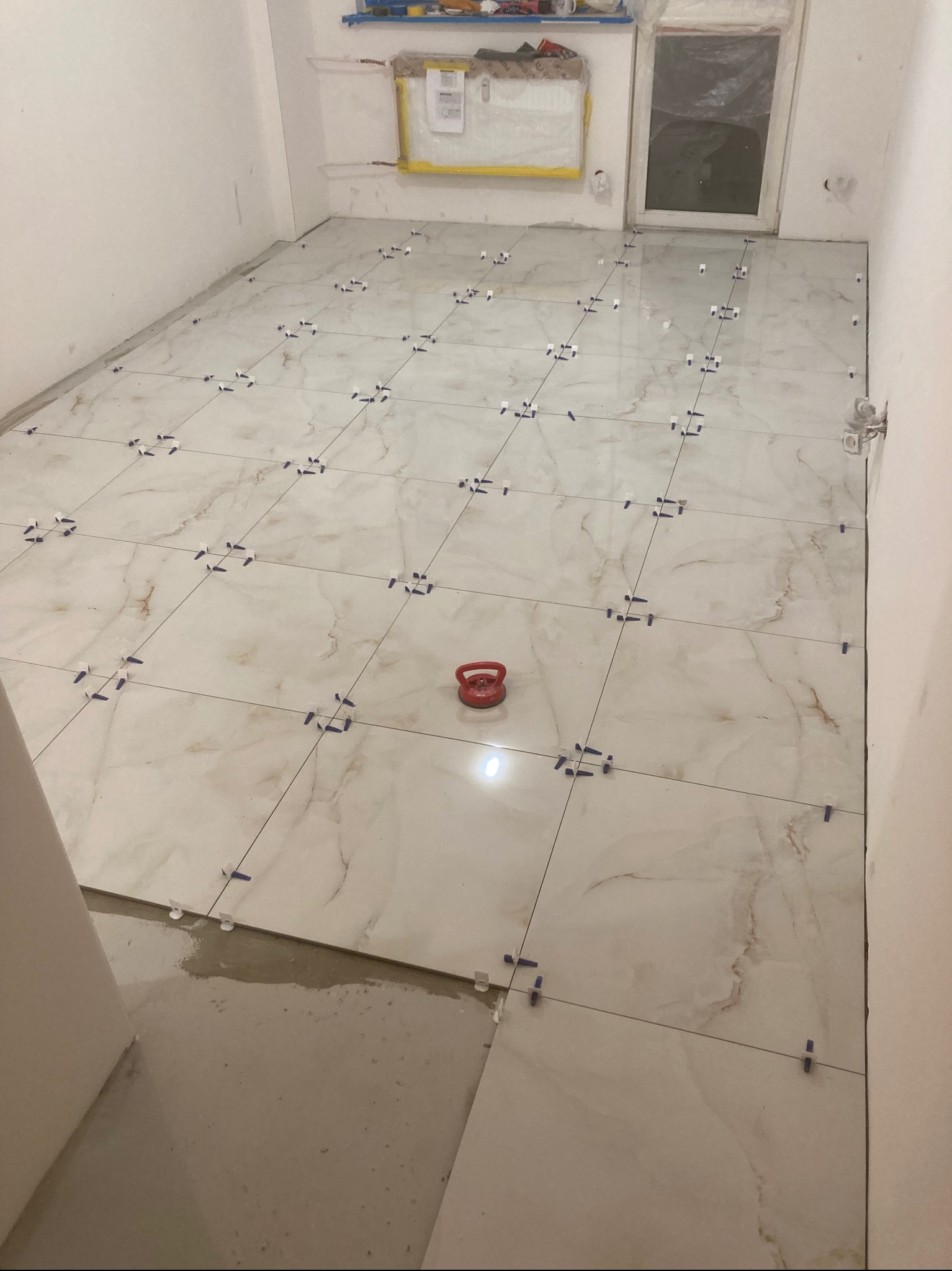 Układanie płytek podłogowych w łazience z użyciem krzyżyków dystansowych i przyssawki. Płytki imitujące marmur, jasne, z brązowymi żyłami. Widoczny fragment wylewki.