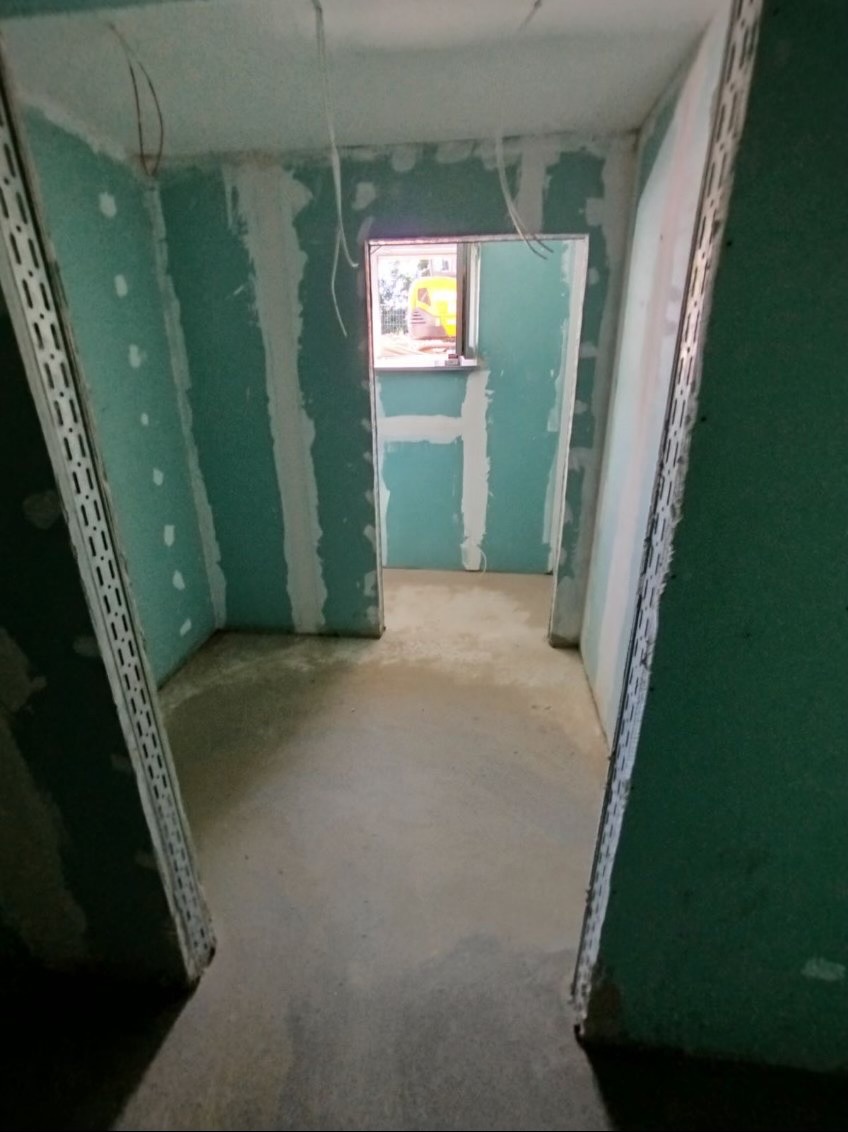 Wnętrze w trakcie prac: ściany z płyt gipsowo-kartonowych, widoczne profile i wstępne szpachlowanie. Surowy beton na podłodze, okno w tle.