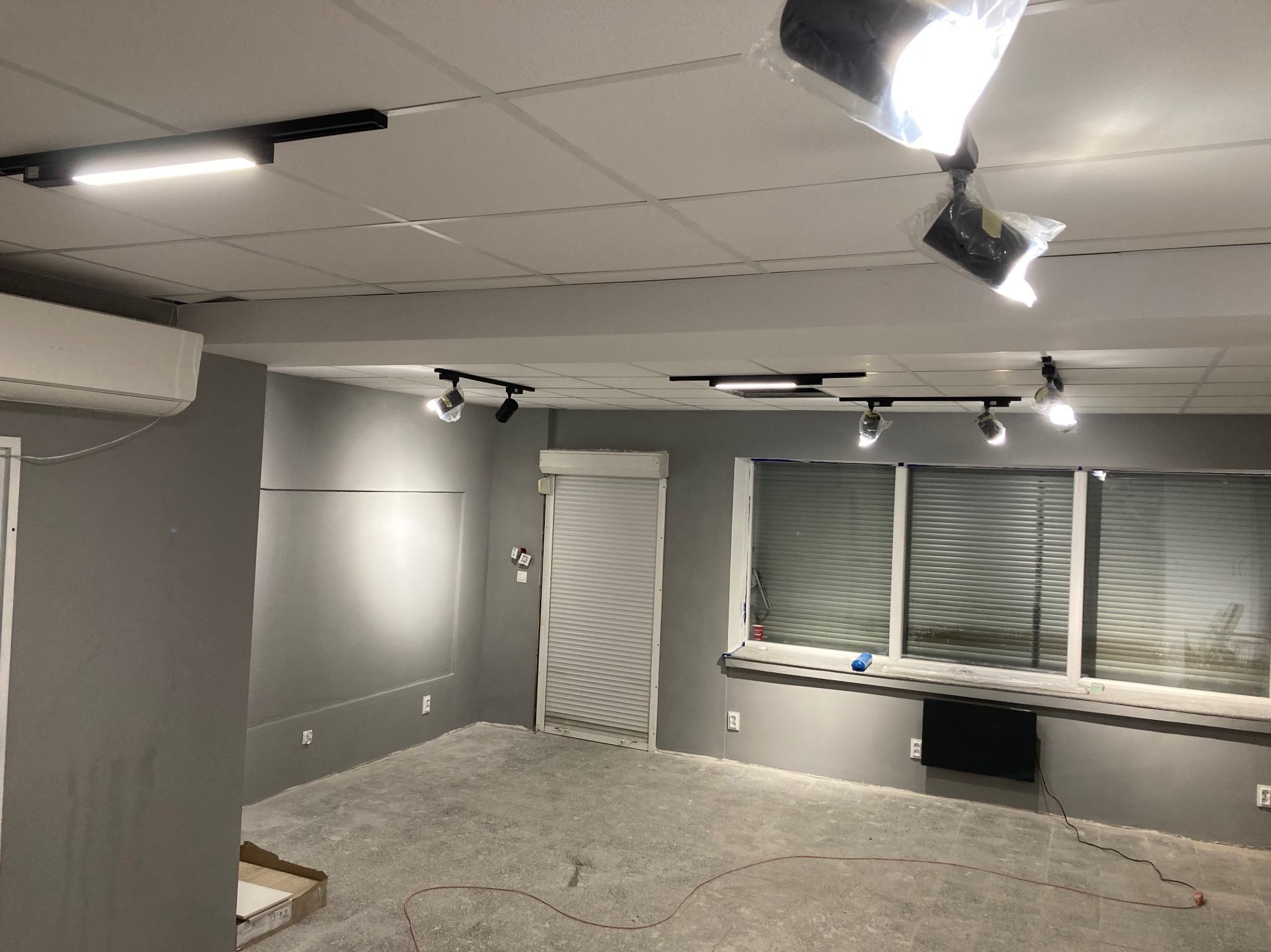 Szare wnętrze biurowe w trakcie remontu: surowy beton na podłodze, ściany pomalowane na szaro, zamontowane oświetlenie szynowe i klimatyzacja. Widoczne okno z żaluzjami.