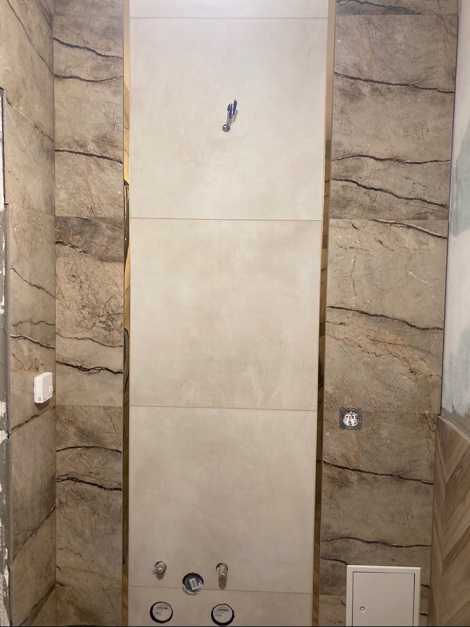 Wykończona ściana w łazience z płytkami imitującymi marmur po bokach i gładkimi, jasnymi płytkami na środku. Widoczne przyłącza wodne i elementy instalacji.