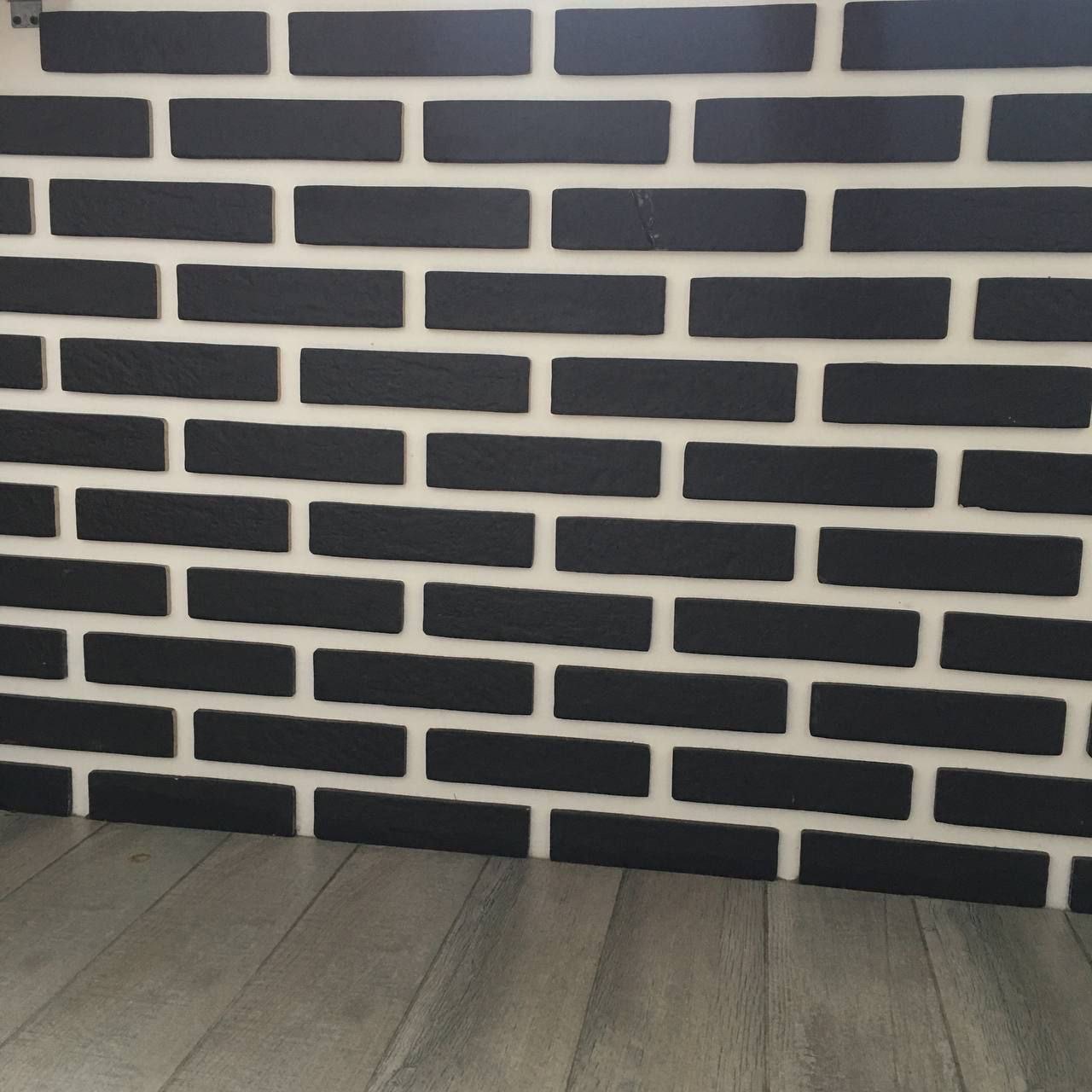 Ściana z czarnych, matowych cegiełek na białej fudze, ułożonych rzędami. Na dole widoczna podłoga z paneli imitujących drewno. Minimalistyczny design.
