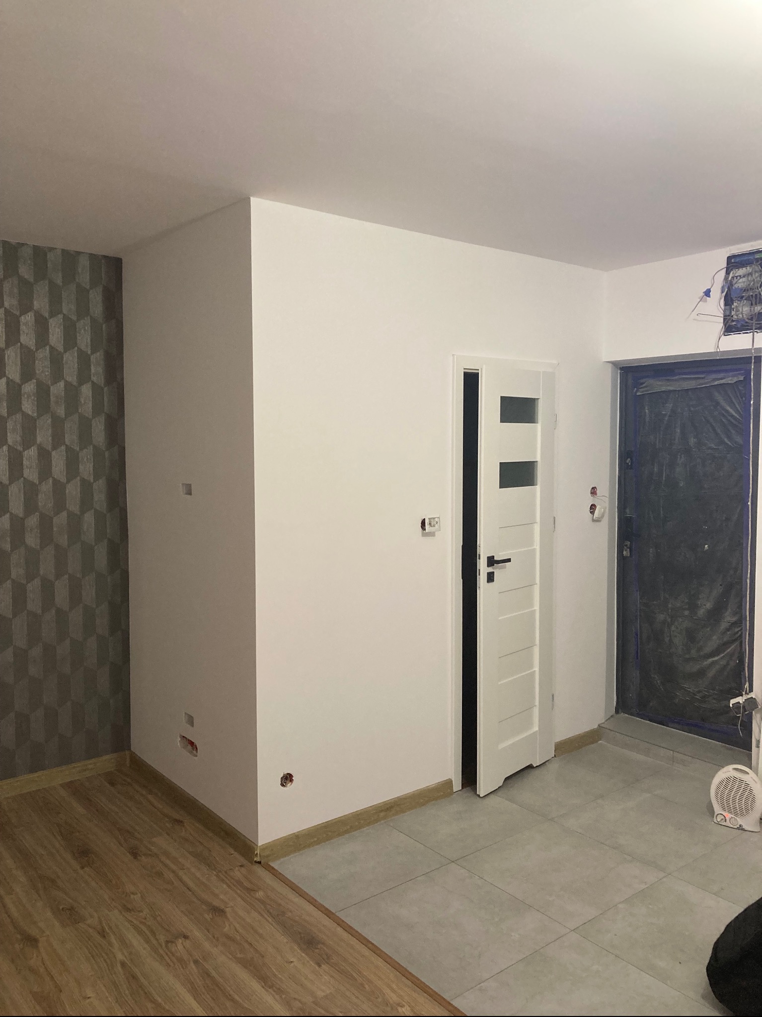 Wnętrze po remoncie: białe ściany, wzorzysta tapeta, drewniana podłoga przechodząca w szare płytki, białe drzwi z przeszkleniami i zabezpieczone folią drzwi wejściowe.