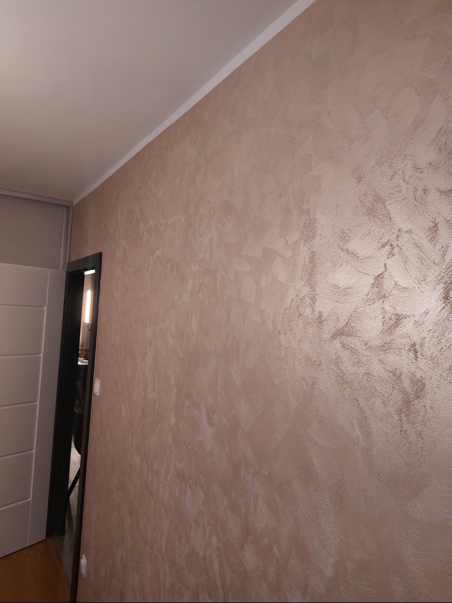 Beżowa ściana z dekoracyjną strukturą, widoczna z perspektywy narożnika pokoju, z fragmentem drzwi po lewej stronie. Subtelne oświetlenie podkreśla fakturę.