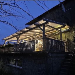 DomLabsStudio - Oświetlona drewniana konstrukcja z przezroczystym zadaszeniem, tworząca zadaszony taras na piętrze domu z kamienną elewacją, widok wieczorny.