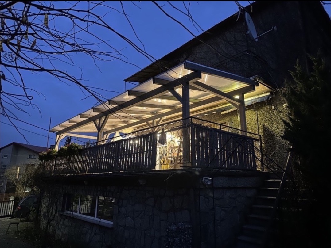 Oświetlona drewniana konstrukcja z przezroczystym zadaszeniem, tworząca zadaszony taras na piętrze domu z kamienną elewacją, widok wieczorny.