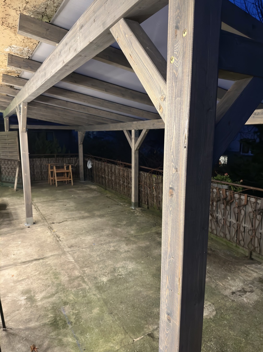 Szara, drewniana konstrukcja z zadaszeniem na betonowym podłożu. Widoczne łączenia belek i słupów. W tle ogrodzenie i zieleń. Ujęcie z dołu, podkreślające solidność konstrukcji.