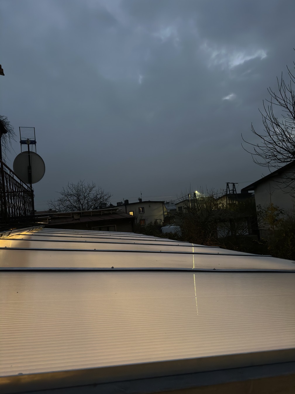 Zadaszenie z poliwęglanu w pochmurny dzień, widok z góry na tle zabudowań i anteny satelitarnej. Delikatne światło odbija się od powierzchni.