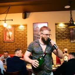 Pavlo LUXO - Mężczyzna z mikrofonem i butelką w dłoni podczas imprezy w Warszawie. W tle siedzący goście i ceglana ściana.