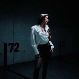 Pavlo LUXO - Kobieta w białej koszuli i czarnej spódnicy pozuje w garażu podziemnym, oświetlona pojedynczą lampą, tworząc dramatyczny efekt światłocienia.