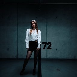 Pavlo LUXO - Kobieta w białej koszuli i czarnej spódnicy pozuje na tle betonowej ściany z numerem 72 w garażu podziemnym, oświetlona pojedynczą lampą.