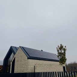 Aura - Dom z panelami słonecznymi na dachu, widoczny zza czarnego ogrodzenia. Kamienna elewacja, nowoczesna architektura i pochmurne niebo w tle. Małe drzewko obok budynku.