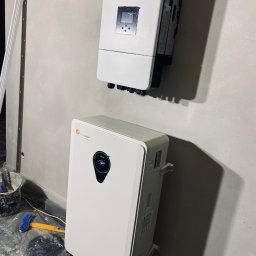 Aura - Biały magazyn energii Felicity Solar i falownik na szarej ścianie. Widoczne narzędzia i wiadro, co sugeruje trwający montaż. Ujęcie z dołu, podkreślające wysokość urządzeń.