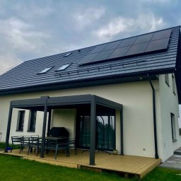 Aura - Nowoczesny dom z panelami fotowoltaicznymi na dachu, pergola z meblami ogrodowymi na tarasie. Biała elewacja, szara dachówka i ciemne panele.