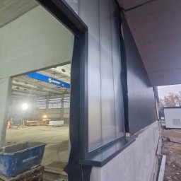 BUDOWNICTWO B.ŻMUDA BŁAŻEJ ŻMUDA - Montaż paneli warstwowych na hali, widok z zewnątrz. Widoczny fragment konstrukcji stalowej i wnętrze hali z suwnicą. Detal łączenia paneli.