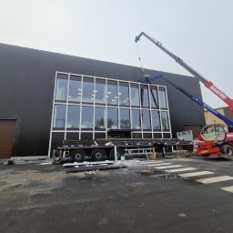 BUDOWNICTWO B.ŻMUDA BŁAŻEJ ŻMUDA - Montaż płyt warstwowych na hali z użyciem dźwigu Manitou. Widoczne płyty na przyczepie i pracownik w koszu dźwigu. Budynek z czarną elewacją i dużymi oknami.