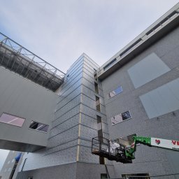 BUDOWNICTWO B.ŻMUDA BŁAŻEJ ŻMUDA - Montaż elewacji hali przemysłowej z podnośnika koszowego. Widoczne płyty warstwowe i elementy konstrukcyjne. Ujęcie z dołu, pochmurne niebo w tle.