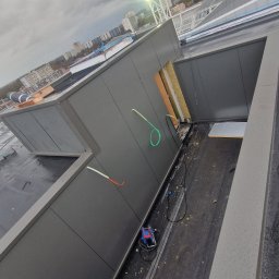 BUDOWNICTWO B.ŻMUDA BŁAŻEJ ŻMUDA - Montaż szarej płyty warstwowej na dachu budynku w trakcie budowy. Widoczny dźwig i elementy konstrukcyjne. Ujęcie z góry, pochmurne niebo w tle.