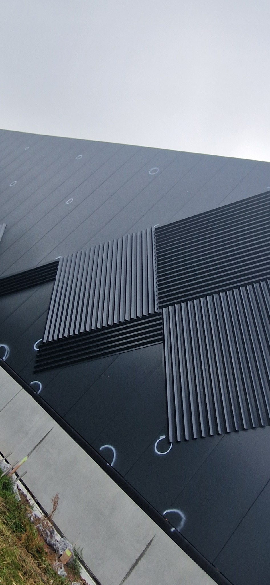 Ciemna elewacja hali z dekoracyjnymi panelami pionowymi i poziomymi, widok z dołu, fragment chodnika i trawy w kadrze. Oznaczenia montażowe widoczne na panelach.