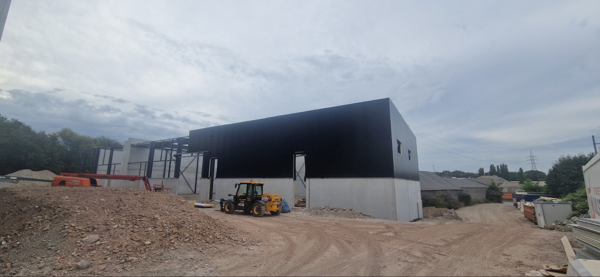 Budowa hali z czarną elewacją z płyt warstwowych, widoczny sprzęt budowlany i konstrukcja stalowa. Ujęcie z ziemi, pochmurne niebo w tle.