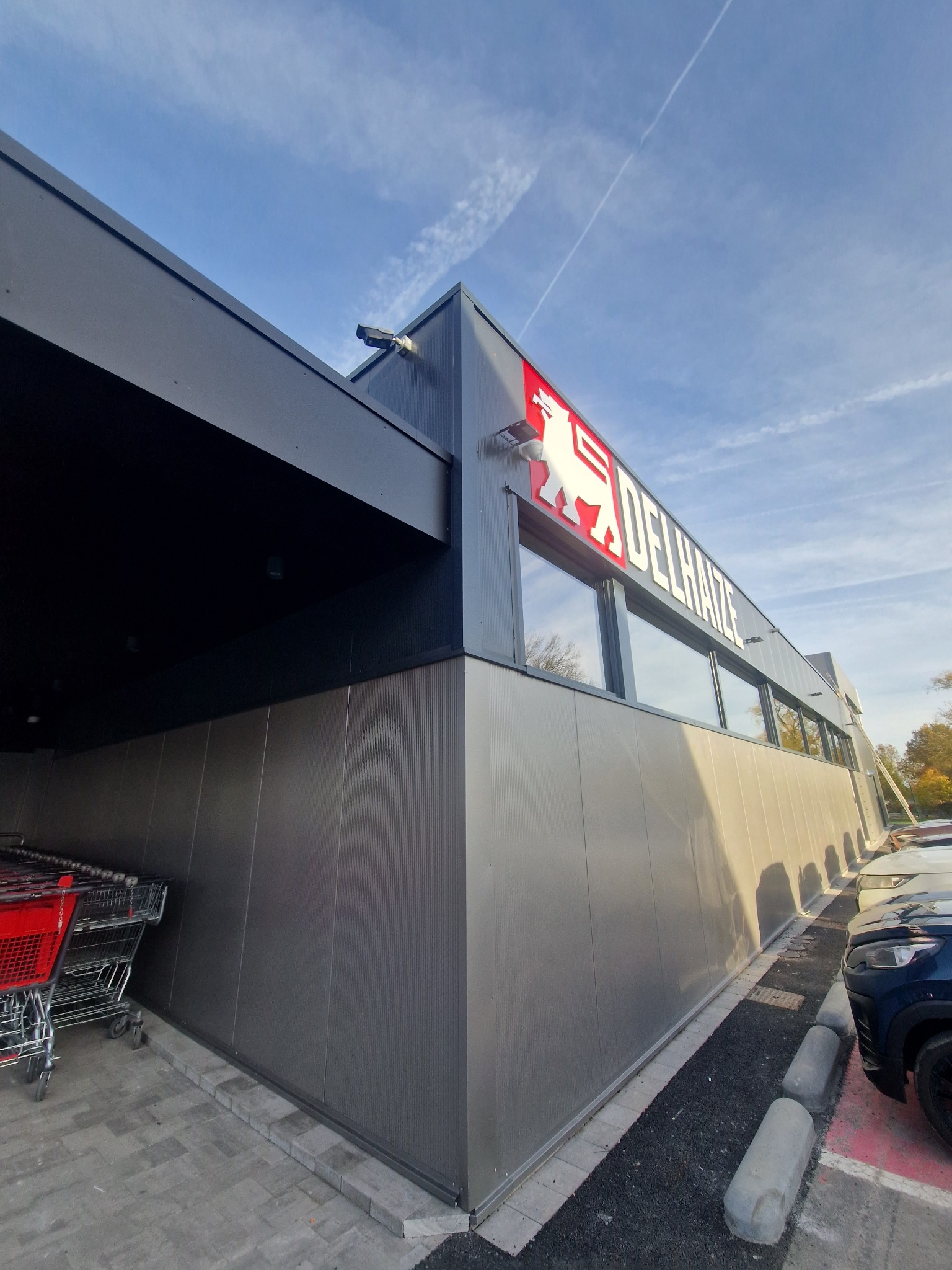 Nowoczesna hala handlowa Delhaize z szarej płyty warstwowej, widok z dołu, błękitne niebo. Wózki sklepowe i auta na parkingu.